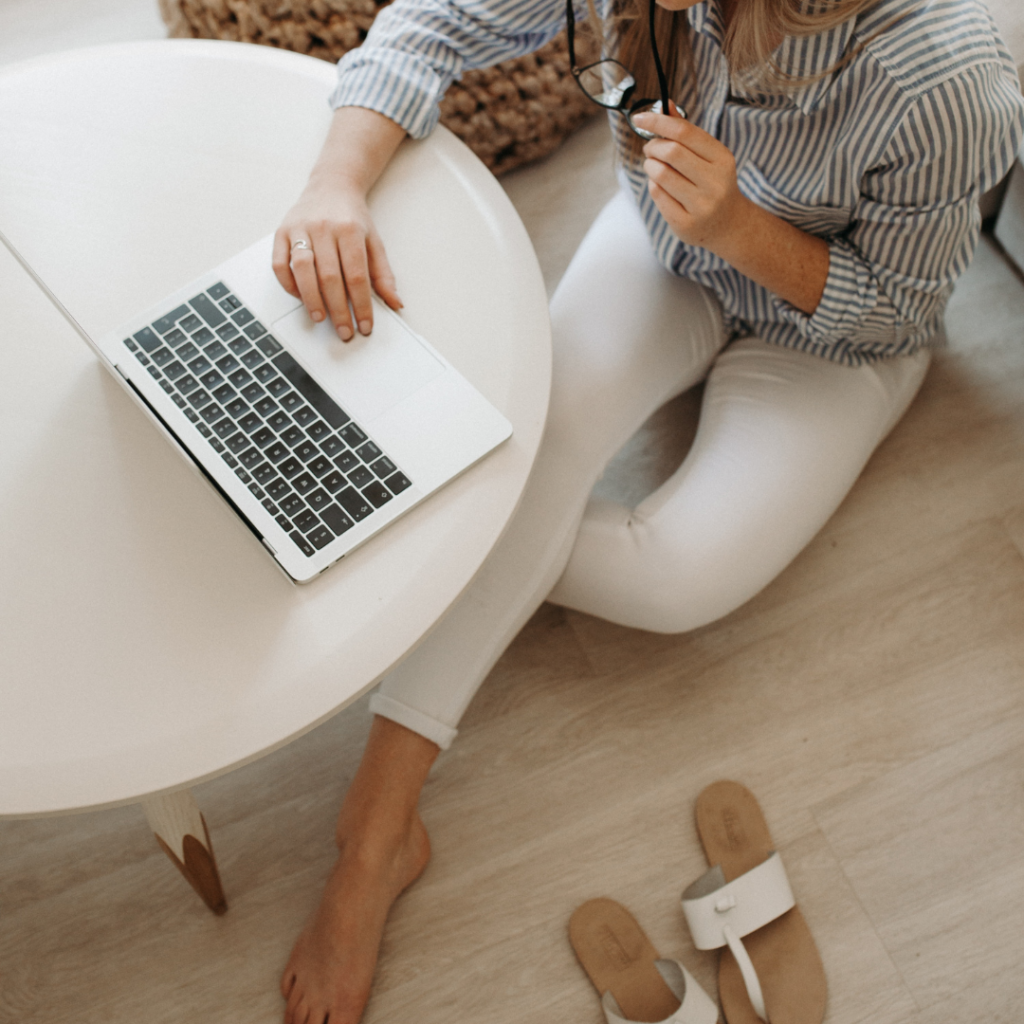 A business woman sitting on the floor, with her shoes off, using her laptop from a bird's eye view. She is working on her handbook from her brand strategist.